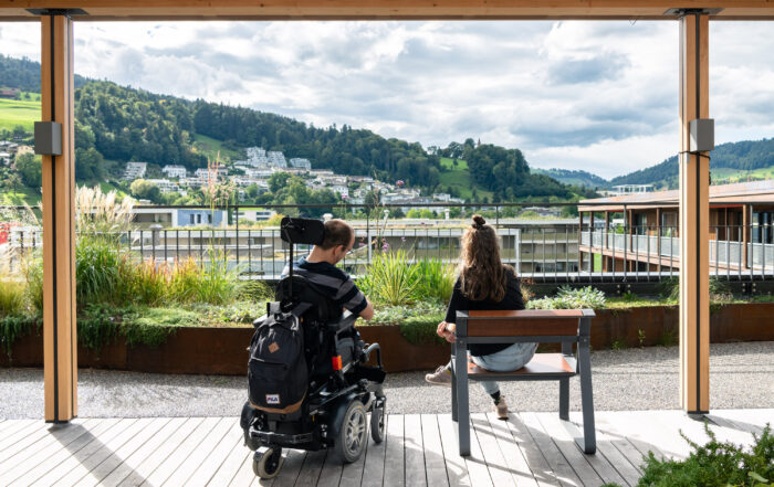 Bild mit Mann im Rollstuhl und Frau auf Holzbank mit Blick vom Balkon