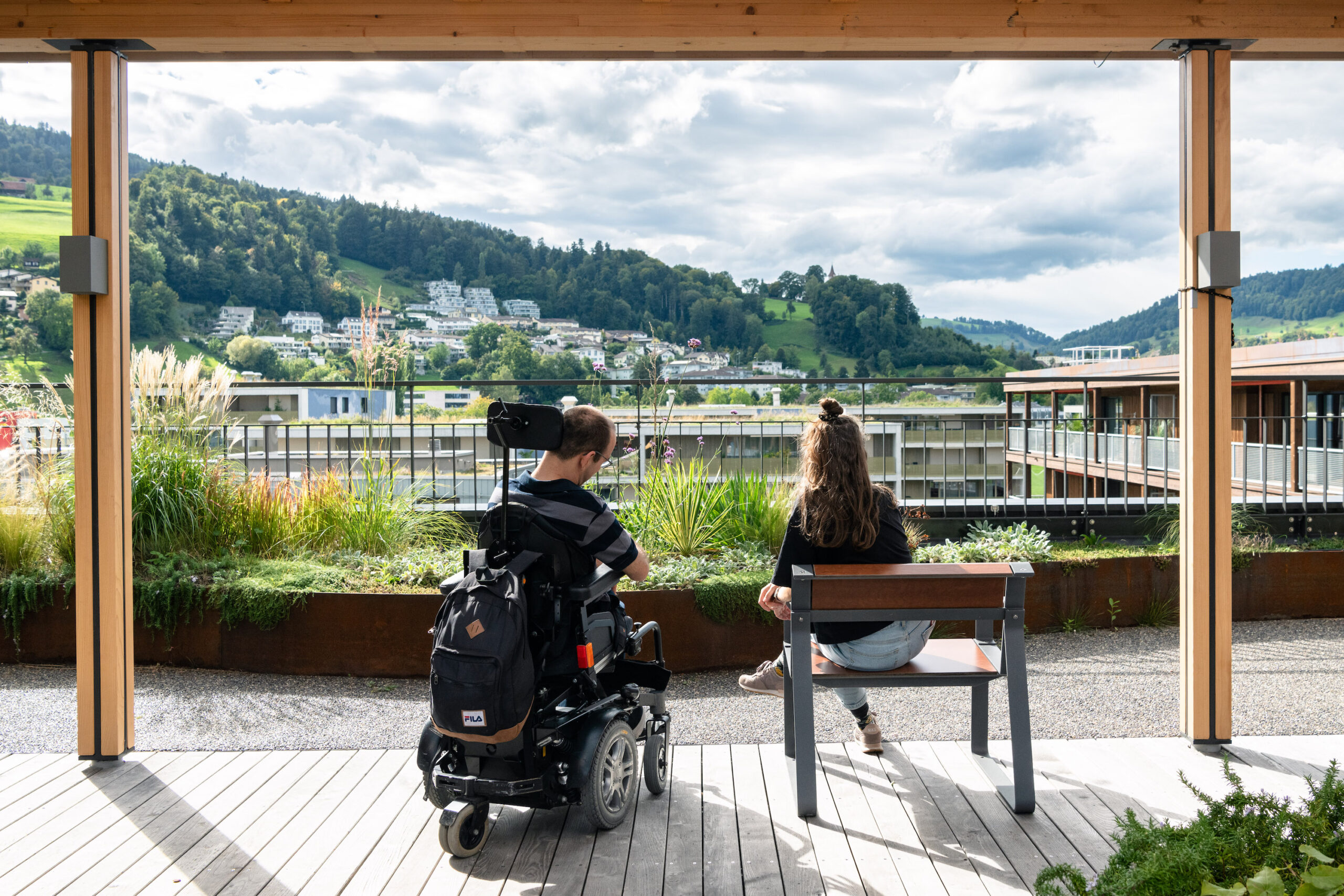 Bild mit Mann im Rollstuhl und Frau auf Holzbank mit Blick vom Balkon